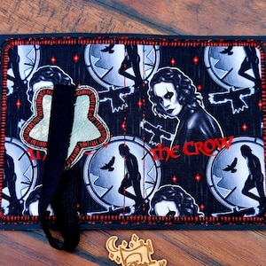May include: A black and red fabric wallet featuring a design from "The Crow" movie. The design includes the character's face, silhouettes, and the movie title in red. A black strap and a clear pen are next to the wallet.