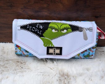 May include: A white zippered wallet with a green Grinch character peeking out from the zipper. The zipper has the words "EW PEOPLE" embroidered on it. The wallet has a colorful pattern on the back.
