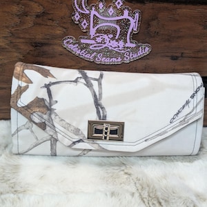 May include: A white and brown camouflage print clutch purse with a gold metal clasp and the text "Conceal Snow" on the front.