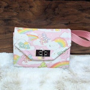 May include: A small, pink and white wallet with a flap closure and a silver clasp. The wallet features a repeating pattern of cartoon characters, rainbows, and clouds. A pink strap is attached to the side. The wallet is on a white, fluffy surface.