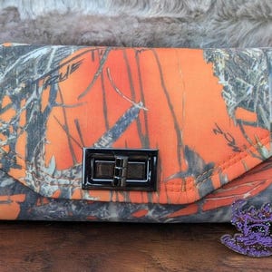 May include: A camouflage print wallet with a bright orange background and a silver clasp. The wallet has a flap closure and a small strap.