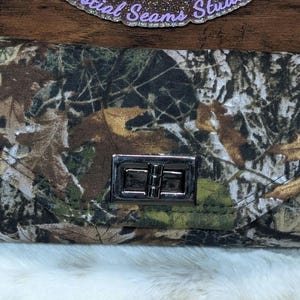 May include: A rectangular wallet with a camouflage pattern featuring shades of green, brown, and tan. The wallet has a silver-colored clasp and a dark brown strap. The background is a wooden surface and white fluffy fabric.