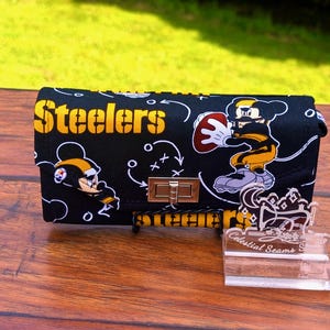May include: Black clutch with gold hardware featuring the word "Steelers" in yellow. The clutch has a cartoon design of a football player. A clear acrylic stand with the text "Celestial Seams" is in front of the clutch.