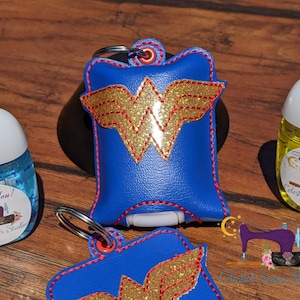 May include: Two blue fabric hand sanitizer holders with a gold glitter Wonder Woman logo. The holders have red stitching and a silver key ring. The holders are on a wooden surface. "Thank You! Celestial Seams Studio" is printed on a label on a clear hand sanitizer bottle.