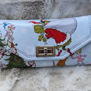 May include: A blue fabric wallet with a Christmas theme featuring the Grinch carrying a bag of presents. The Grinch is green and wearing a red Santa suit. The wallet has a gold clasp and a red and white striped strap.