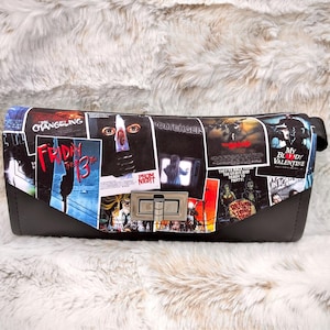 May include: A black clutch featuring a collage of horror movie posters, including "Friday the 13th" and "My Bloody Valentine." The bag has a silver clasp and a black base. The background is a fluffy, white surface.