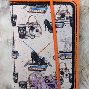 May include: A white and black patterned fabric notebook cover with a black and orange trim. The cover features a repeating pattern of coffee cups, high heels, purses, and glasses. The notebook cover has a black strap and a black and orange bookmark.
