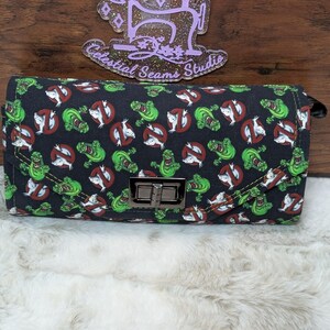 May include: A black clutch wallet with a repeating pattern of green ghosts and red no-ghost symbols. The wallet has a silver clasp and a black zipper pull. The Celestial Seams Studio logo is visible in the background.