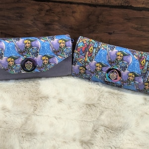 May include: Two rectangular wallets featuring a repeating pattern of a blonde character in a green tunic holding a sword and shield against a blue and purple background. Each wallet has a central circular clasp with a shield emblem design.