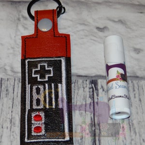 May include: A red leather keychain with a black and white embroidered image of a vintage video game controller. The keychain has a silver button and a black metal ring. The keychain is next to a white lip balm tube with a purple and white design.