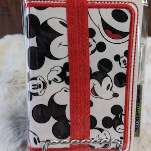 May include: A small notebook featuring a repeating pattern of Mickey Mouse faces in black on a white background. The notebook is bound with a red elastic band and has a red embroidered border. A pen is included.