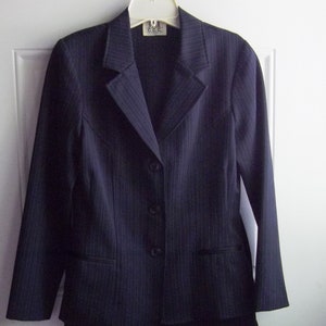May include: A navy blue pinstripe suit, consisting of a blazer and a skirt, hangs on a white hanger against a white door. The blazer has a notched lapel, three buttons, and two front pockets. The skirt is a straight, knee-length style.