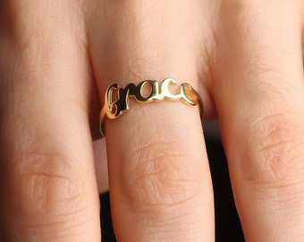 Op de afbeelding: Een goudkleurige ring met de naam "Grace" in een sierlijk lettertype. De ring wordt aan een vinger gedragen, met een vage achtergrond. De ring is een delicaat sieraad.