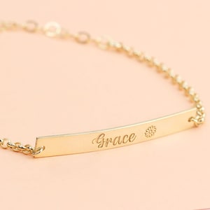 Könnte beinhalten: Ein goldenes Kettenarmband mit einem kleinen rechteckigen Anhänger, auf dem das Wort "Grace" und ein kleines Blumenmuster eingraviert sind.