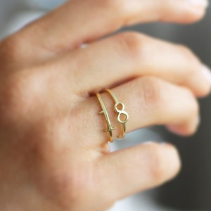 May include: A hand wearing three gold rings. One ring features a cross, another has the number eight, and the third is a simple band. The rings are thin and delicate, with a polished finish. The background is blurred.