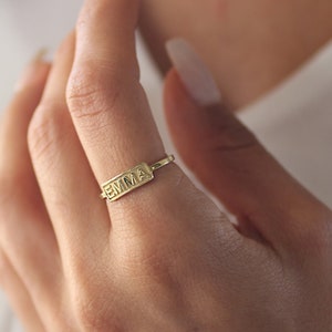 May include: A gold ring featuring the name "EMMA" inscribed on a rectangular plaque. The ring is worn on a finger, set against a white top. This is a piece of jewellery.