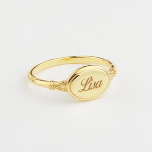 May include: A gold signet ring with the name "Lisa" engraved on the oval face. The ring has a delicate band and decorative detailing on the shoulders. The ring is set against a white background.