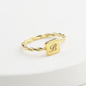 Op de afbeelding: Een goudkleurige ring met een gedraaide band en een vierkant middelpunt. Het vierkant heeft de letter "B" in een sierlijk lettertype. De ring ligt op een witte ondergrond.