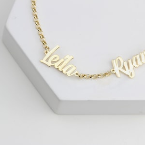 May include: A gold-colored necklace with the names "Leila" and "Ryan" in a cursive font. The names are connected by a delicate chain. The necklace is displayed on a white, geometric surface.