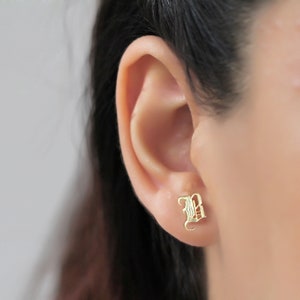 May include: Gold initial letter 'B' stud earrings. The earrings are in a gothic font style. The earrings are shown in a person's ear.