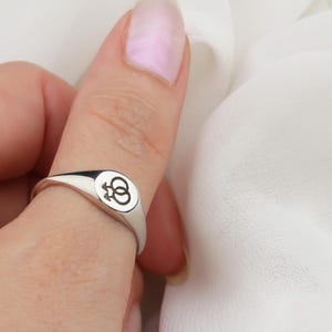Può includere: Un anello chevalier in argento con una faccia rettangolare arrotondata. La faccia dell'anello presenta un simbolo in bianco e nero. L'anello è indossato su un dito. Lo sfondo è un tessuto bianco.
