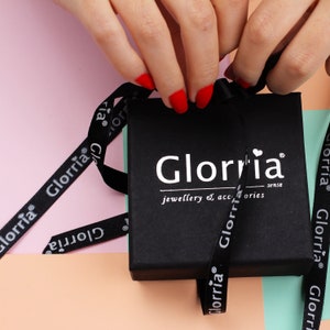 May include: A black jewelry box with the brand name "Glorria" in white lettering. The box is tied with a black ribbon printed with the brand name. The background is a mix of pink, peach, and green.
