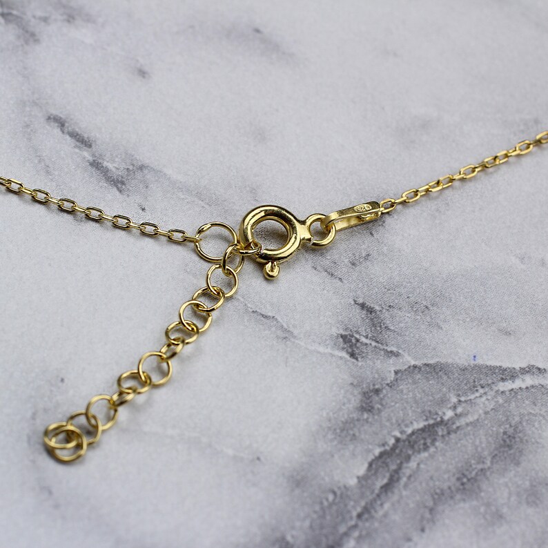 K&ouml;nnte beinhalten: Nahaufnahme eines goldfarbenen Halskettenverschlusses und einer Kette. Die Halskette hat eine filigrane Kette mit einem Karabinerverschluss und einer verstellbaren Verl&auml;ngerungskette. Das Metall hat eine helle, gl&auml;nzende Oberfl&auml;che. Der Hintergrund ist ein hellgraues und wei&szlig;es Marmormuster.