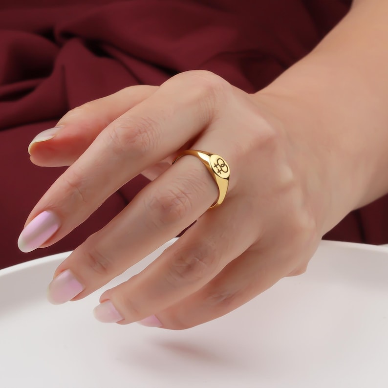 May include: A gold-colored ring with a rounded face featuring a small, black symbol. The ring is worn on a finger, with the hand resting against a white surface and a burgundy background.