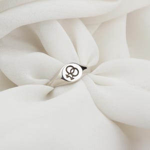 Puede incluir: Un anillo de sello plateado con una cara circular con un s&iacute;mbolo femenino doble negro. El anillo est&aacute; sobre un fondo blanco texturizado.