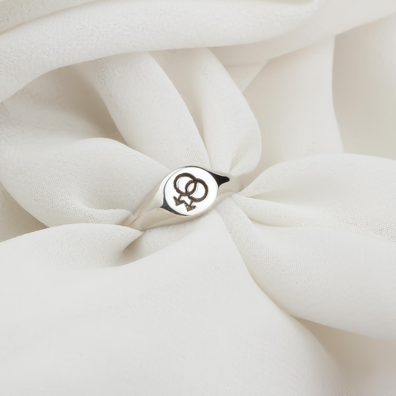 Puede incluir: Un anillo de sello plateado con una cara circular con un s&iacute;mbolo femenino doble negro. El anillo est&aacute; sobre un fondo blanco texturizado.