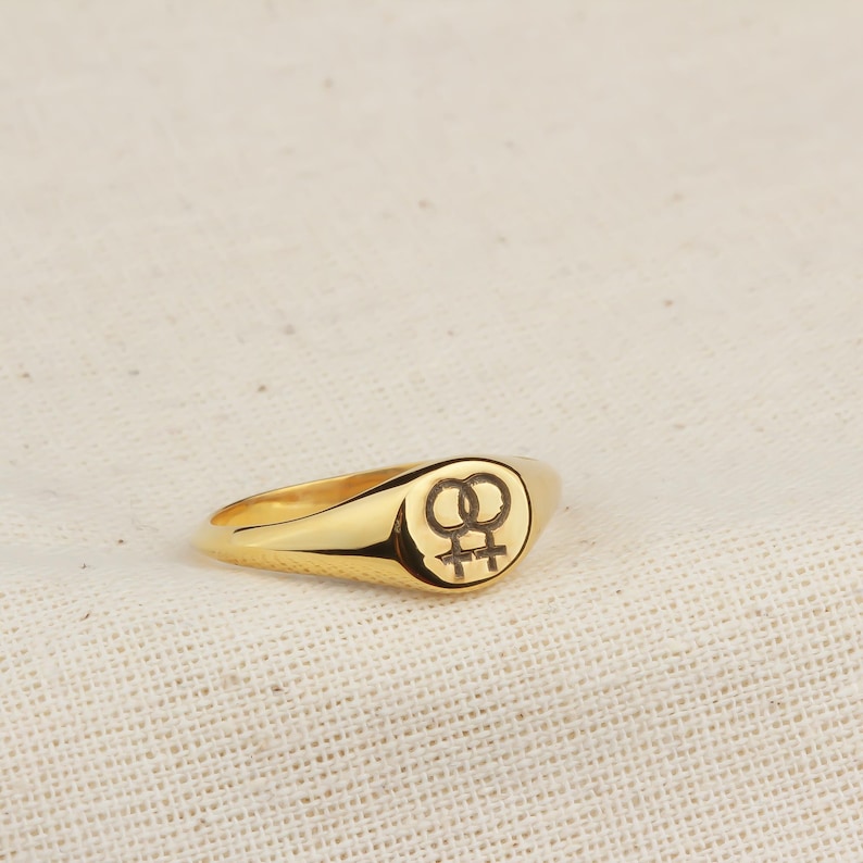 Puede incluir: Un anillo de sello dorado con una cara redondeada. El anillo presenta un s&iacute;mbolo femenino doble negro estilizado. El anillo est&aacute; sobre un fondo neutro y texturizado.