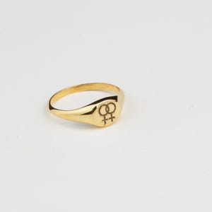 Puede incluir: Un anillo de sello dorado con una superficie lisa y pulida. El anillo presenta una cara rectangular redondeada con un s&iacute;mbolo negro grabado. El anillo est&aacute; sobre un fondo blanco.