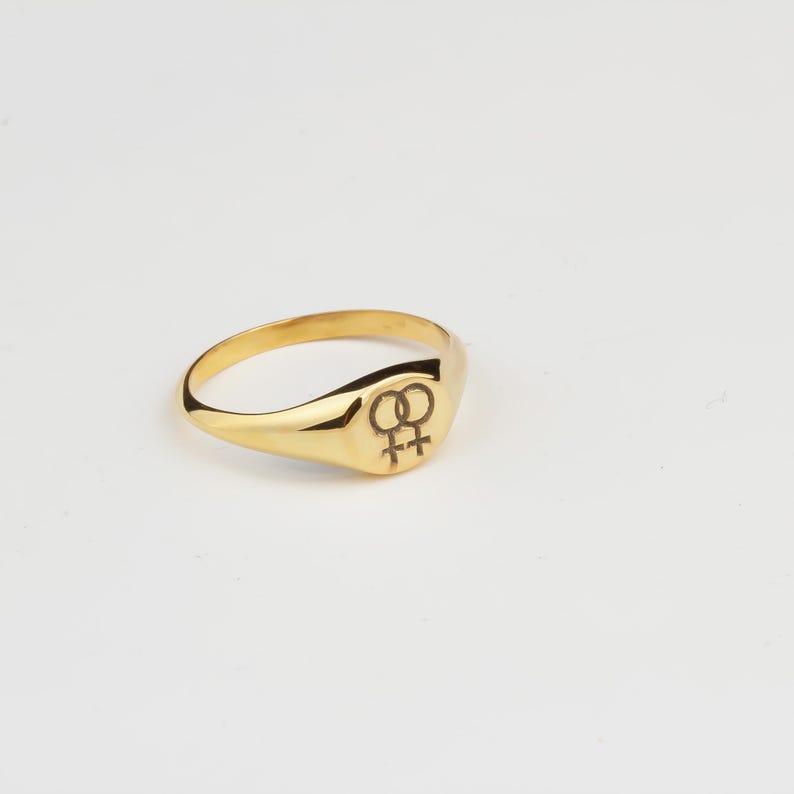 Puede incluir: Un anillo de sello dorado con una superficie lisa y pulida. El anillo presenta una cara rectangular redondeada con un s&iacute;mbolo negro grabado. El anillo est&aacute; sobre un fondo blanco.