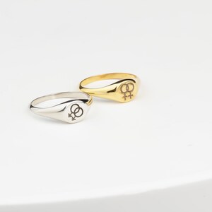 Puede incluir: Dos anillos de sello, uno plateado y otro dorado, cada uno con un s&iacute;mbolo femenino doble. Los anillos se muestran sobre una superficie blanca. El anillo dorado tiene un acabado pulido, mientras que el plateado es mate.