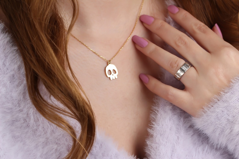 May include: A gold chain necklace with a skull pendant. The necklace is worn by a person with long brown hair and is visible against a light purple fuzzy background.