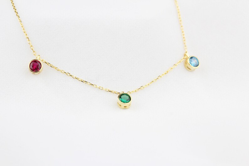 K&ouml;nnte beinhalten: Eine zarte goldene Halskette mit drei gefassten Edelsteinen: einem Rubin, einem Smaragd und einem blauen Topas. Die Edelsteine sind gleichm&auml;&szlig;ig auf der feinen Kette verteilt und bilden ein schlichtes, aber elegantes Design. Die Halskette wird auf wei&szlig;em Hintergrund pr&auml;sentiert.