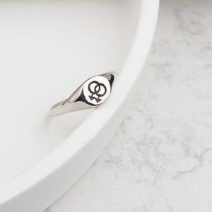 Puede incluir: Un anillo plateado con una cara redonda con un s&iacute;mbolo l&eacute;sbico negro. El anillo est&aacute; sobre una superficie blanca. La banda del anillo es lisa y pulida, reflejando la luz.