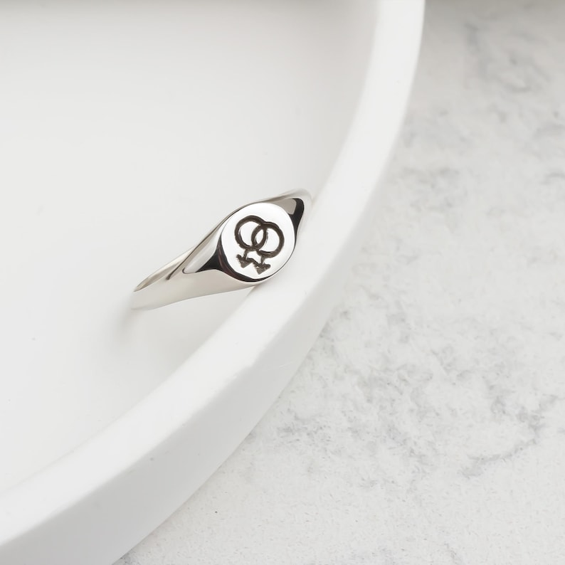 Puede incluir: Un anillo plateado con una cara redonda con un s&iacute;mbolo l&eacute;sbico negro. El anillo est&aacute; sobre una superficie blanca. La banda del anillo es lisa y pulida, reflejando la luz.