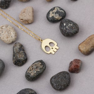 May include: A gold chain necklace with a small, cutout skull pendant. The necklace is displayed on a background of small, grey and brown stones.