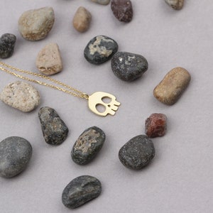 May include: A gold chain necklace with a delicate skull pendant. The necklace is displayed on a grey surface with small stones.
