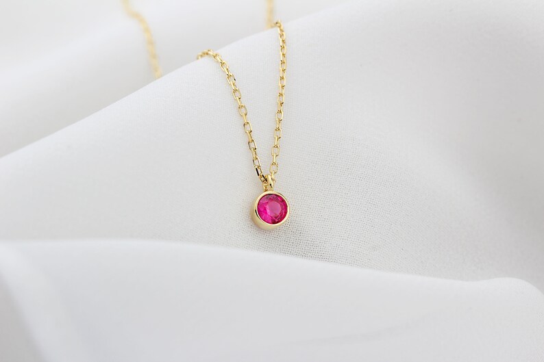 May include: A delicate gold chain necklace with a small, round, bright pink gemstone pendant.