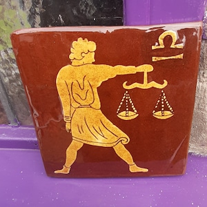 May include: A brown ceramic tile featuring a gold illustration of the astrological sign Libra. The illustration shows a figure holding a scale with two pans.