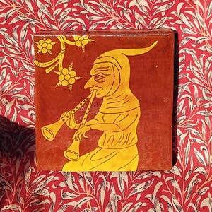 May include: A square ceramic tile featuring a deep red background with a yellow illustration of a figure in a pointed hood playing a double-piped wind instrument. The design includes stylised yellow floral motifs in the upper left corner.