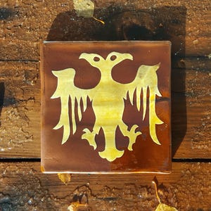 May include: Square brown tile featuring a gold double-headed eagle design. The eagle is centrally placed and has a detailed, stylized appearance. The tile is set against a weathered wooden surface.