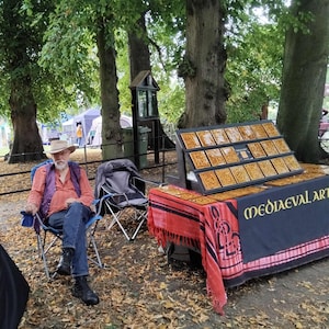 May include: An outdoor display of art pieces with a black table cloth that reads "MEDIEVAL ART". The art pieces are square, with a gold-colored design. A person is sitting in a blue chair in front of the display.
