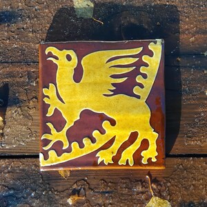 May include: Square ceramic tile featuring a yellow griffin design on a maroon background. The griffin, a mythical creature, has a lion's body and an eagle's head and wings. The tile is likely for decorative use, possibly for a wall or floor.