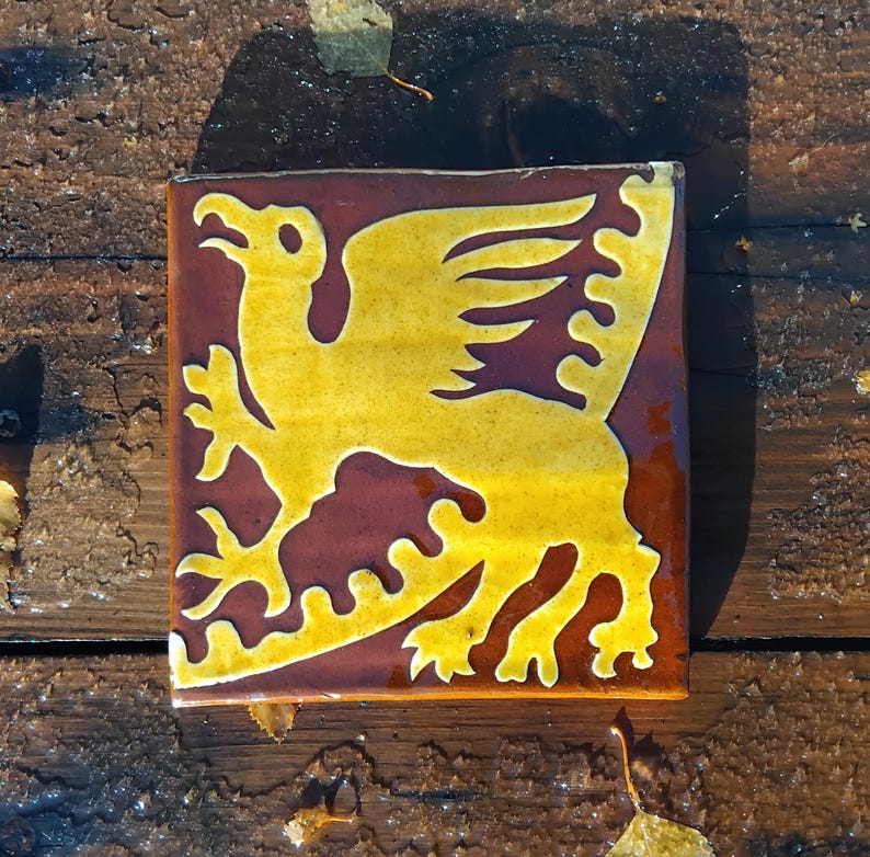May include: Square ceramic tile with a yellow griffin design on a maroon background. The griffin is a mythical creature with the body of a lion and the head and wings of an eagle. The tile is likely for decorative use.