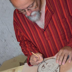 May include: A person wearing a red and orange striped shirt with glasses is carving a design into a clay tile. The tile features a circular pattern with a central animal figure. The artist is using a small tool to create the intricate details.