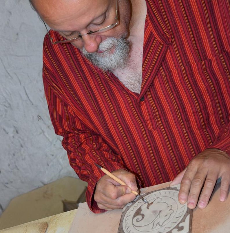 May include: A person wearing a red and orange striped shirt with glasses is carving a design into a clay tile. The tile features a circular pattern with a central animal figure. The artist is using a small tool to create the intricate details.