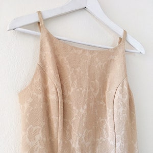May include: A sleeveless, beige dress with a floral pattern. The dress is hanging on a white hanger.